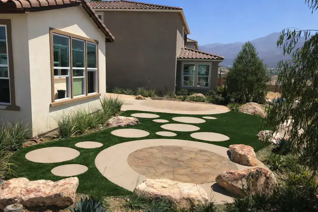 A backyard with mountain views, featuring various sizes of circular concrete stepping stones and stone patios in a lush artificial turf lawn.