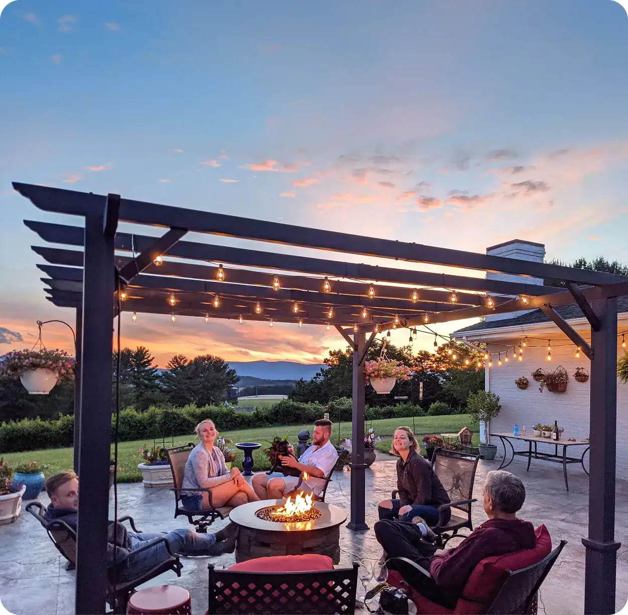 Family and friends relaxing around a fire pit under a pergola with string lights at sunset