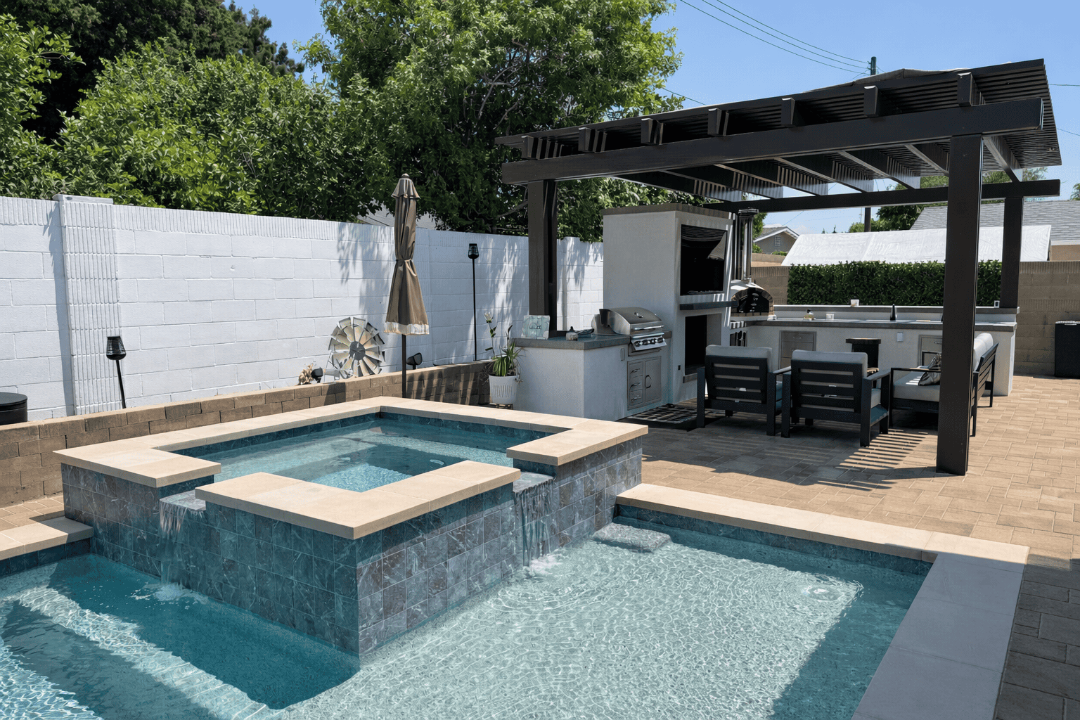 Modern backyard pool and spa with waterfall feature, outdoor kitchen, pergola, and lounge seating.