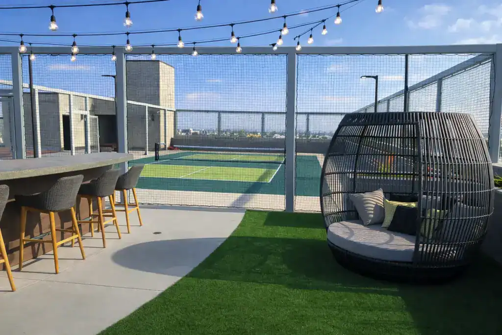 A commercial rooftop patio showing artificial turf for lounging and a fenced-in sports court for pickleball or tennis in the background.