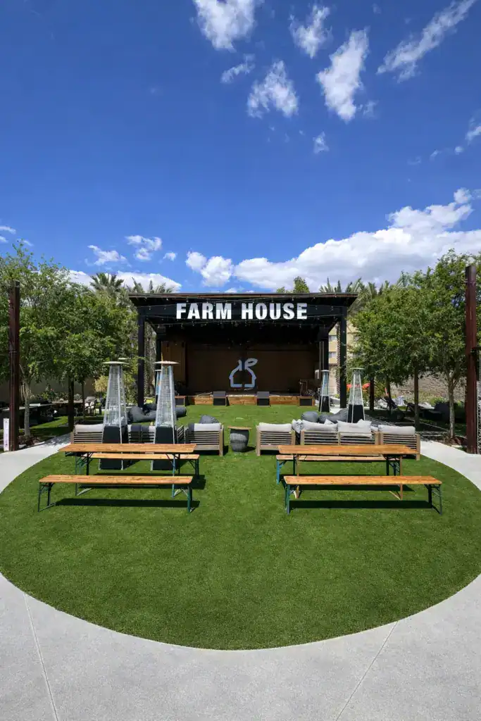 A large artificial turf event lawn at an outdoor venue featuring a stage, picnic tables, and patio heaters under a bright blue sky.