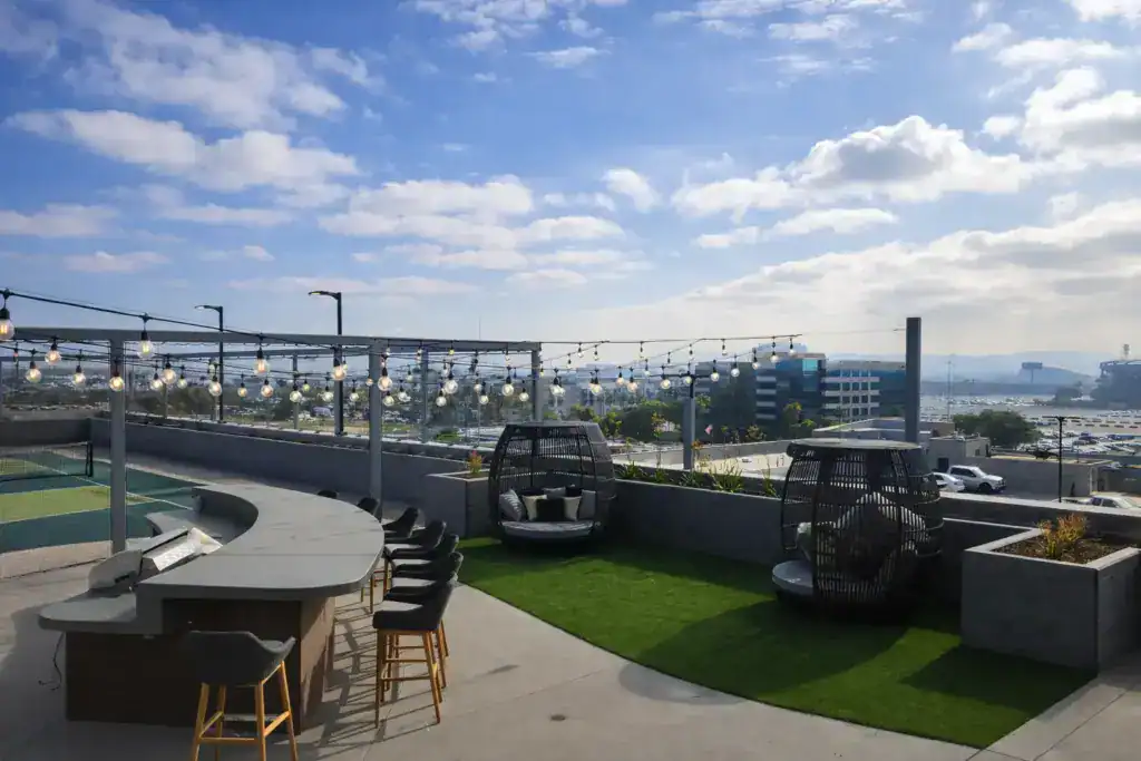 A luxury rooftop lounge featuring an outdoor bar, wicker daybeds, string lights, and a vibrant artificial turf area